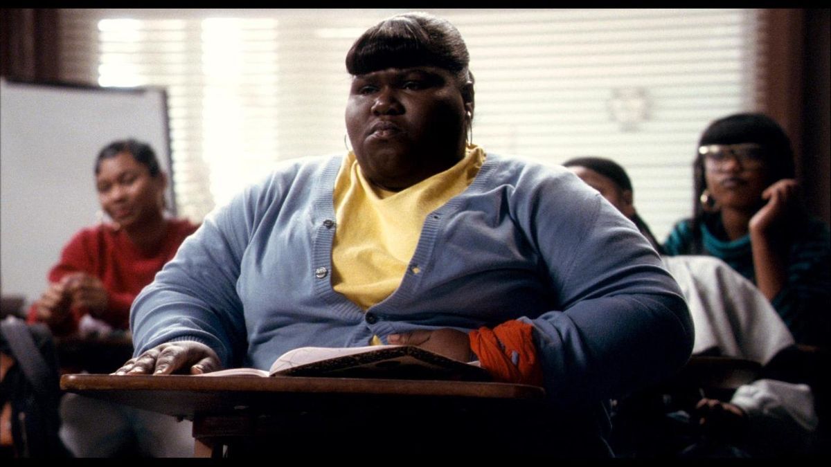 Cortés Filmworks: A person wearing a light blue cardigan and a yellow shirt sits at a wooden desk with an open book in a classroom, as though captured by Cortés Filmworks. They are looking forward with a serious expression, capturing the precious moment of focused learning. In the background, three other people are visible, sitting and chatting casually, providing an authentic backdrop for this beautifully documented scene. : CortesFilmworks.com